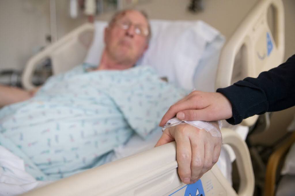 Elderly man is in a hospital bed with his hand stretched out and someone has their hand of his